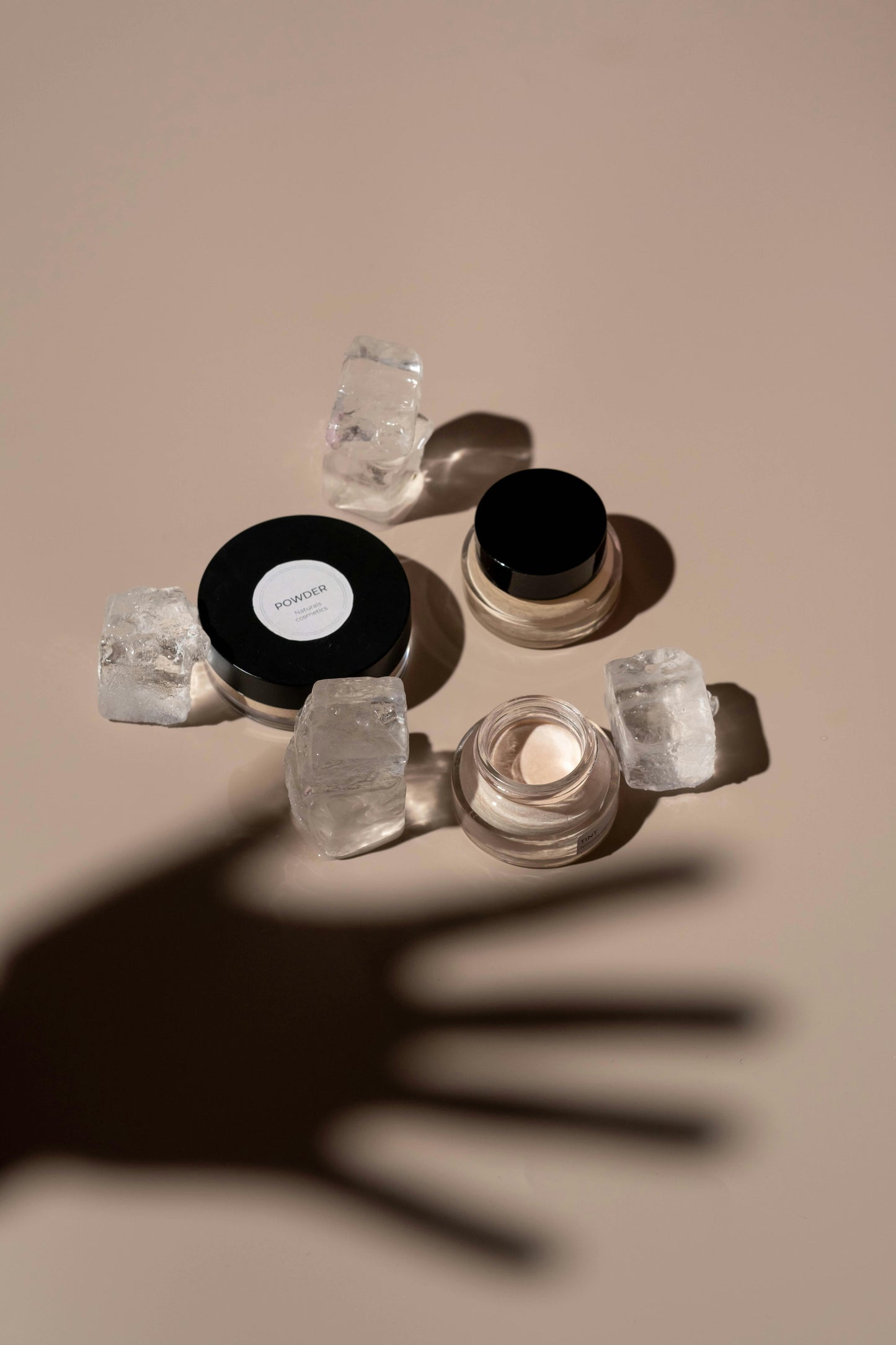 Minimalist skincare jars and powder container placed with ice cubes on beige surface, casting shadow of an outstretched hand.