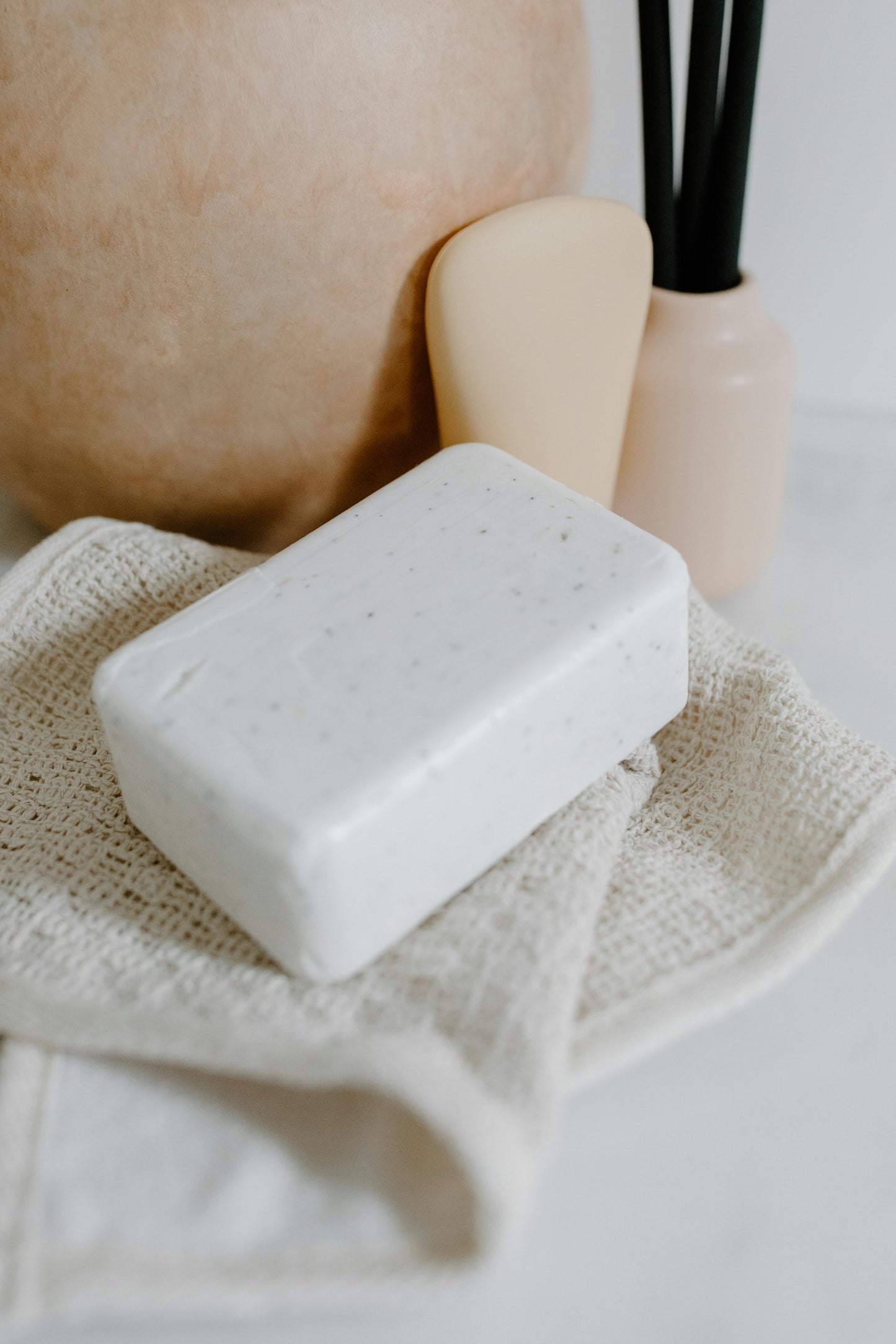 Rectangular white soap brick resting on a textured beige cloth with a beige scraper and soft-toned decor in the background.