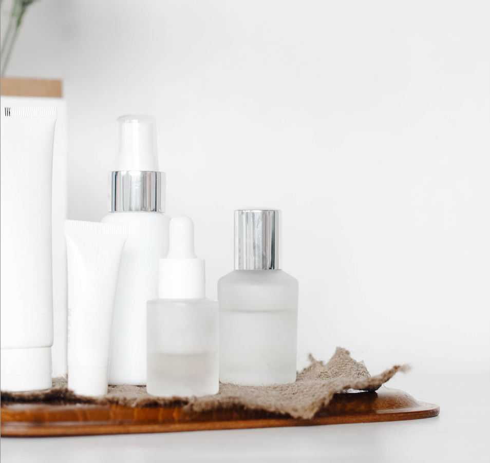 Assortment of white and frosted glass skincare bottles and tubes arranged on a wooden tray with textured cloth base.