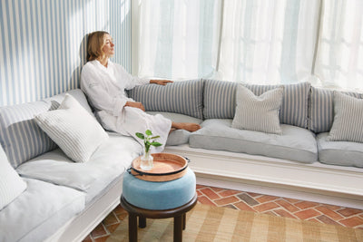 A woman in a white bathrobe relaxing on a grey sofa with soft white curtains in the background, capturing a serene moment of self-care with Sommetbeauty.