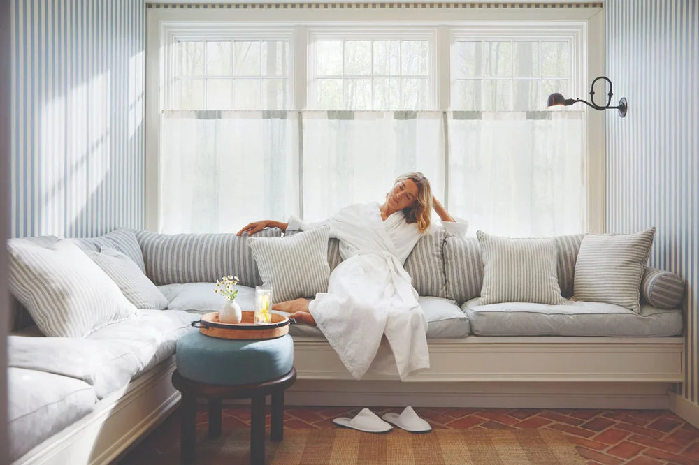 A woman in a white bathrobe relaxing on a grey sofa with soft white curtains in the background, capturing a serene moment of self-care with Sommetbeauty.