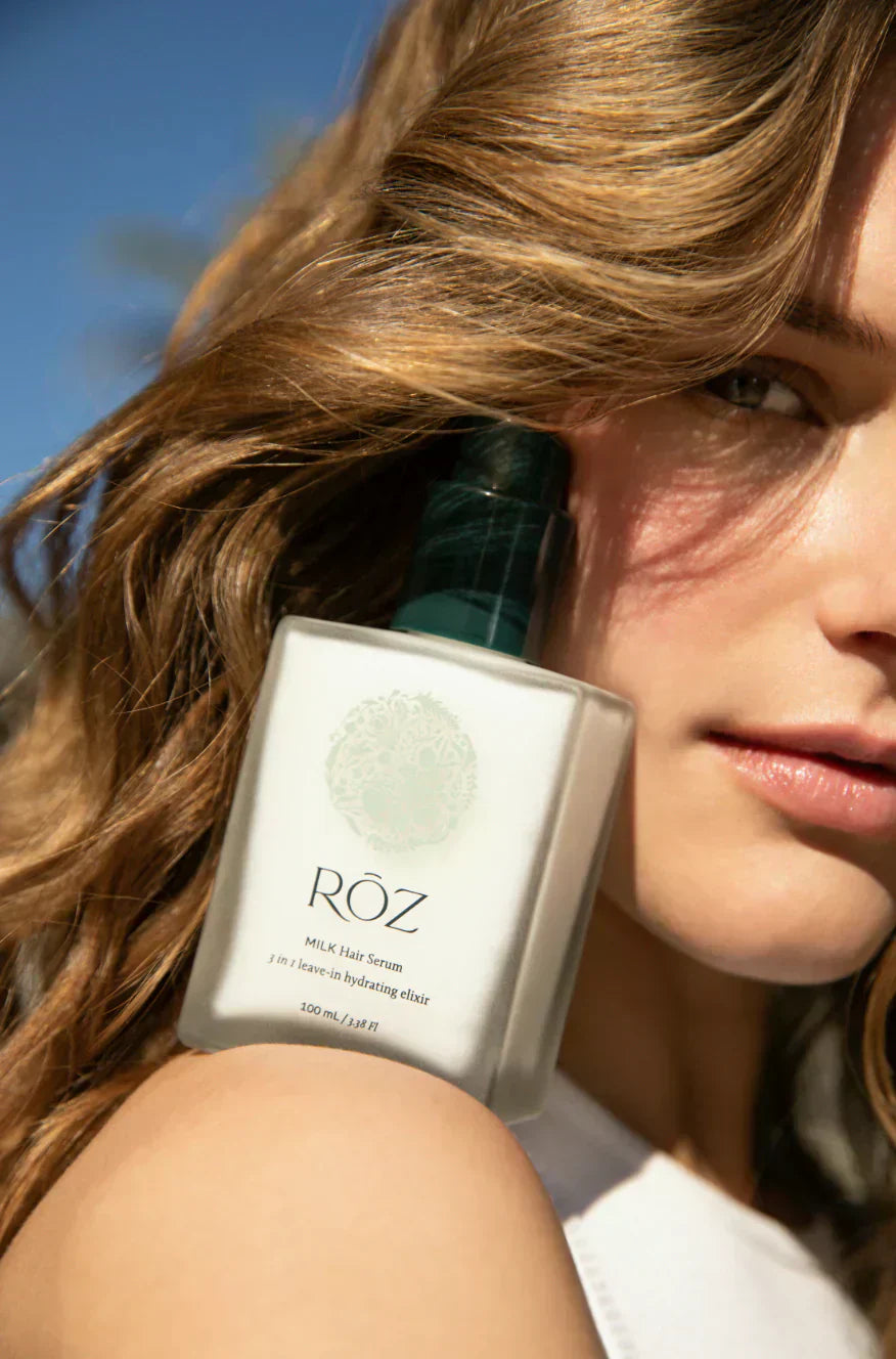 Close-up of a woman holding Sommetbeauty’ RŌZ MILK Hair Serum with her wavy hair in focus.